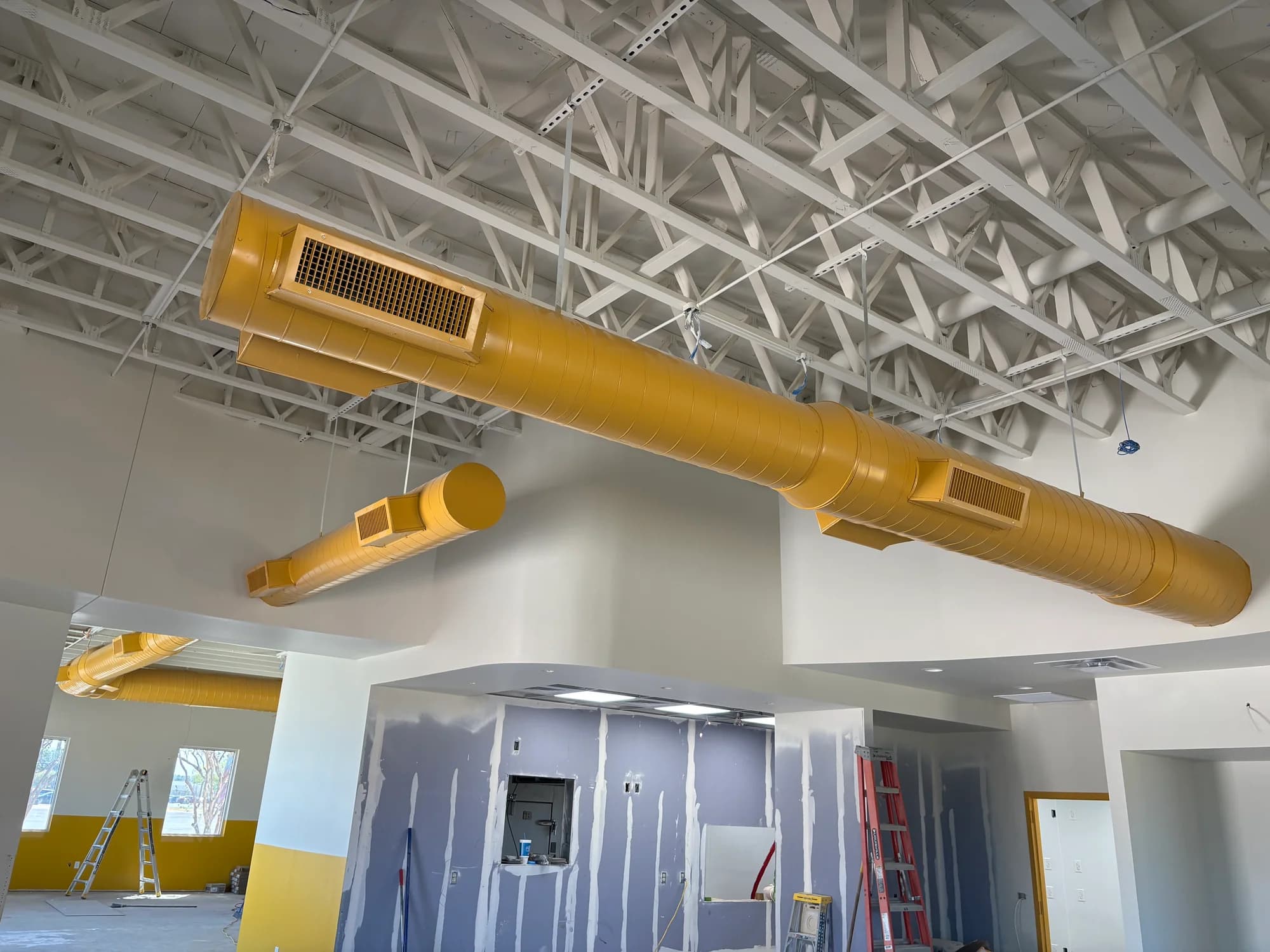 Yellow exposed spiral ductwork and matching grilles beneath white roof trusses at Wienerschnitzel in Woodway